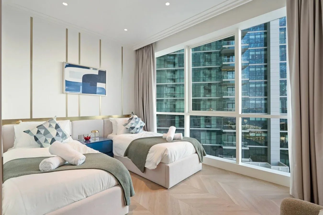 Un dormitorio luminoso con dos camas junto a grandes ventanas con vistas a edificios cercanos.