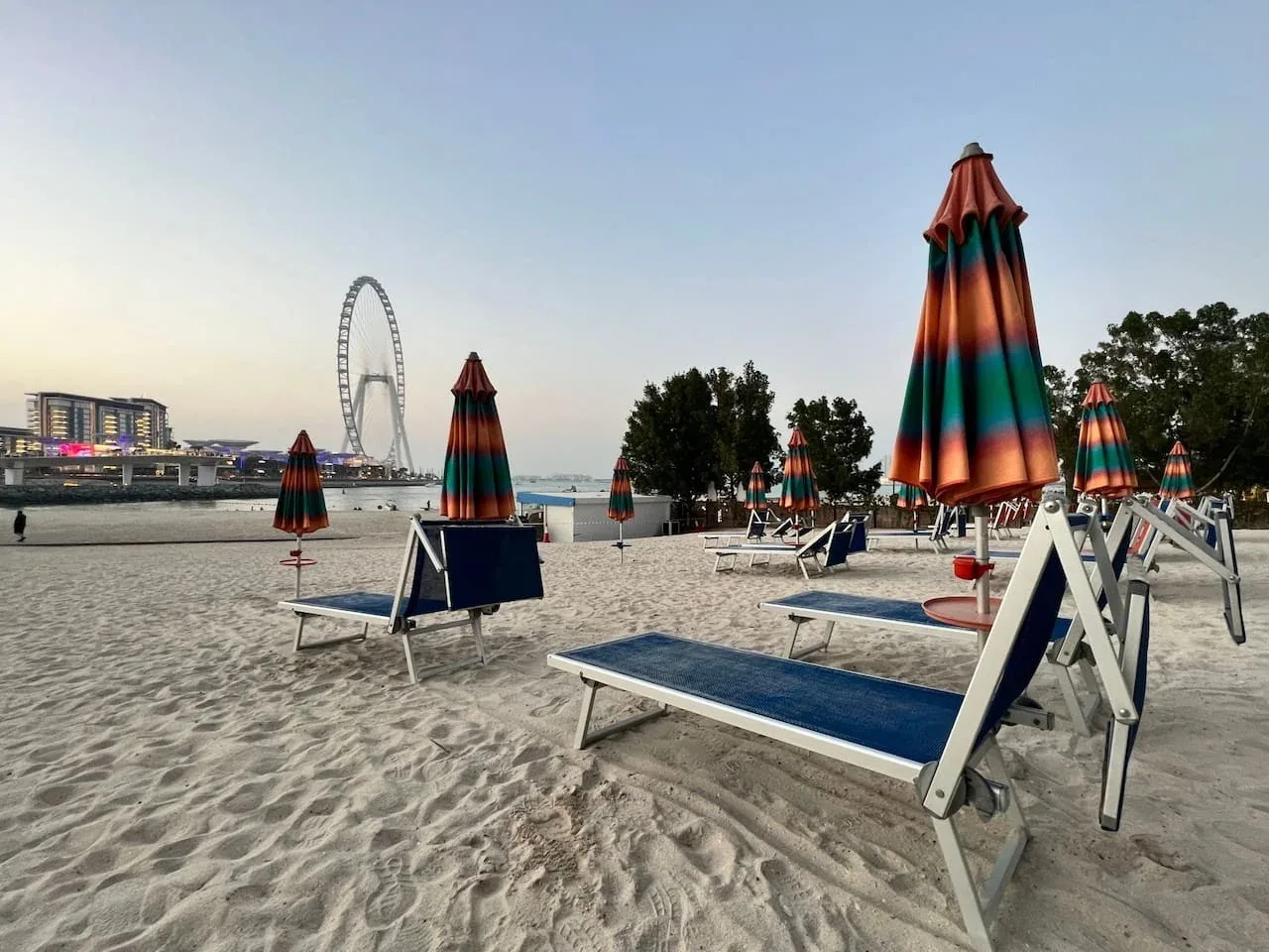 Una playa de arena con tumbonas y sombrillas de colores cerradas, y una gran noria a lo lejos.