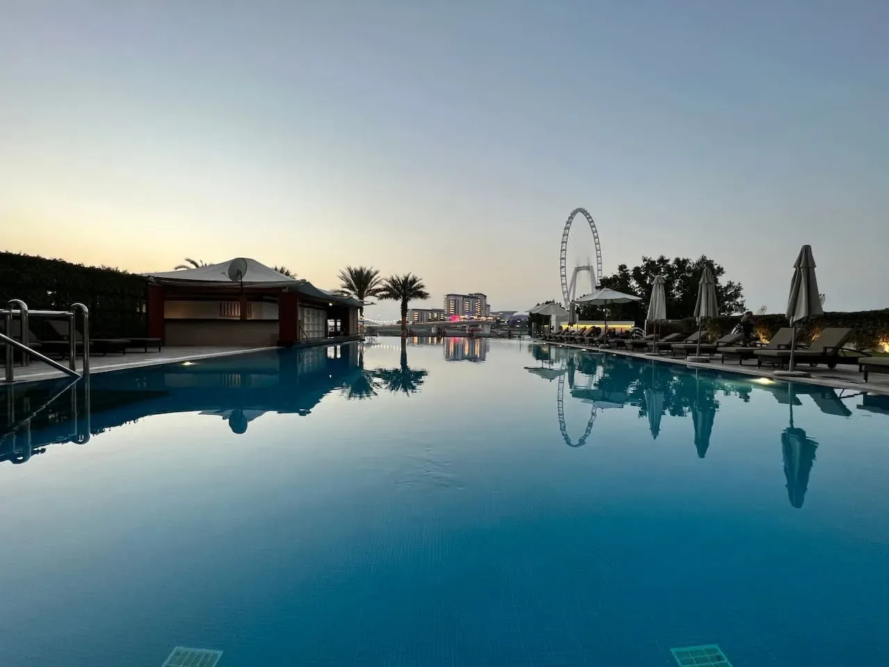 Una piscina al aire libre refleja palmeras y una gran noria al anochecer.