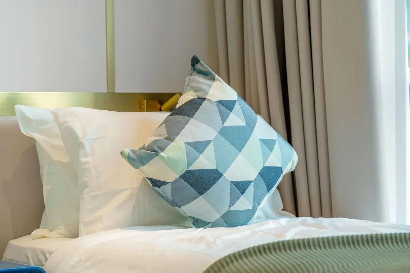 Una cama de dormitorio está tendida con ropa de cama blanca y un cojín geométrico.