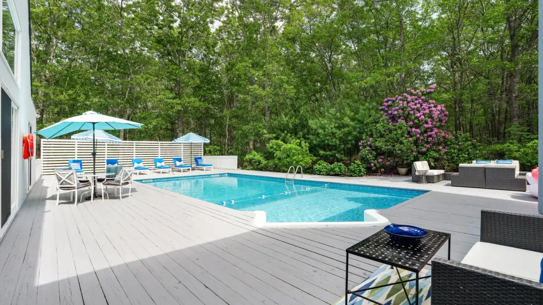 A backyard pool sits beside a spacious deck, with trees and outdoor seating.