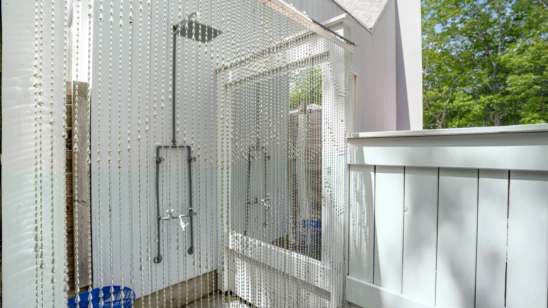 A modern outdoor shower area with a glass enclosure and leafy trees beyond.
