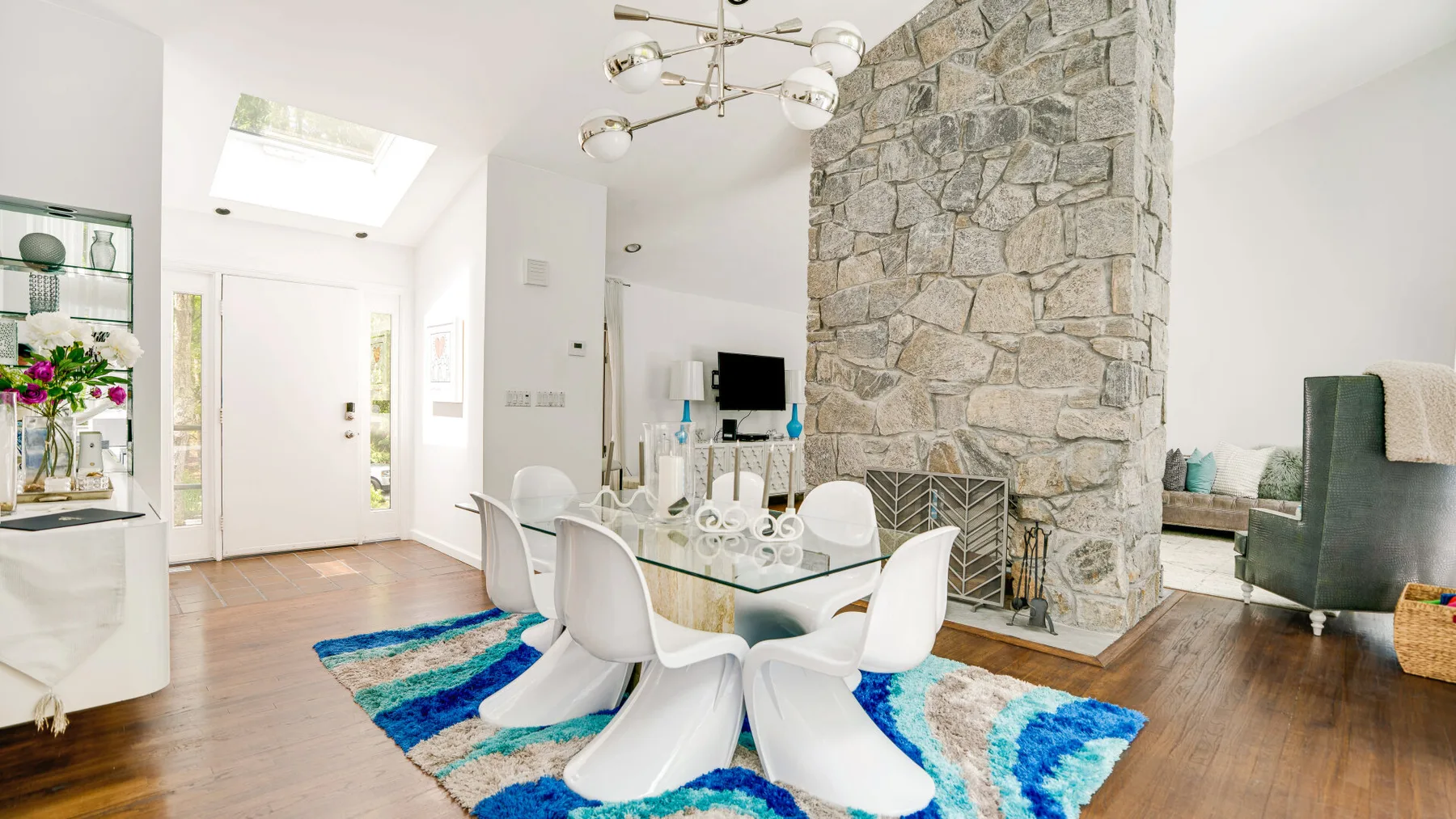 A bright dining area has a glass table beside a large stone fireplace.