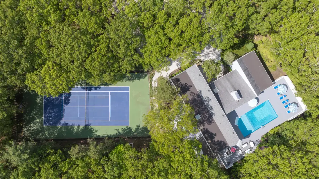 An aerial view shows a tennis court beside a house and swimming pool, surrounded by trees.