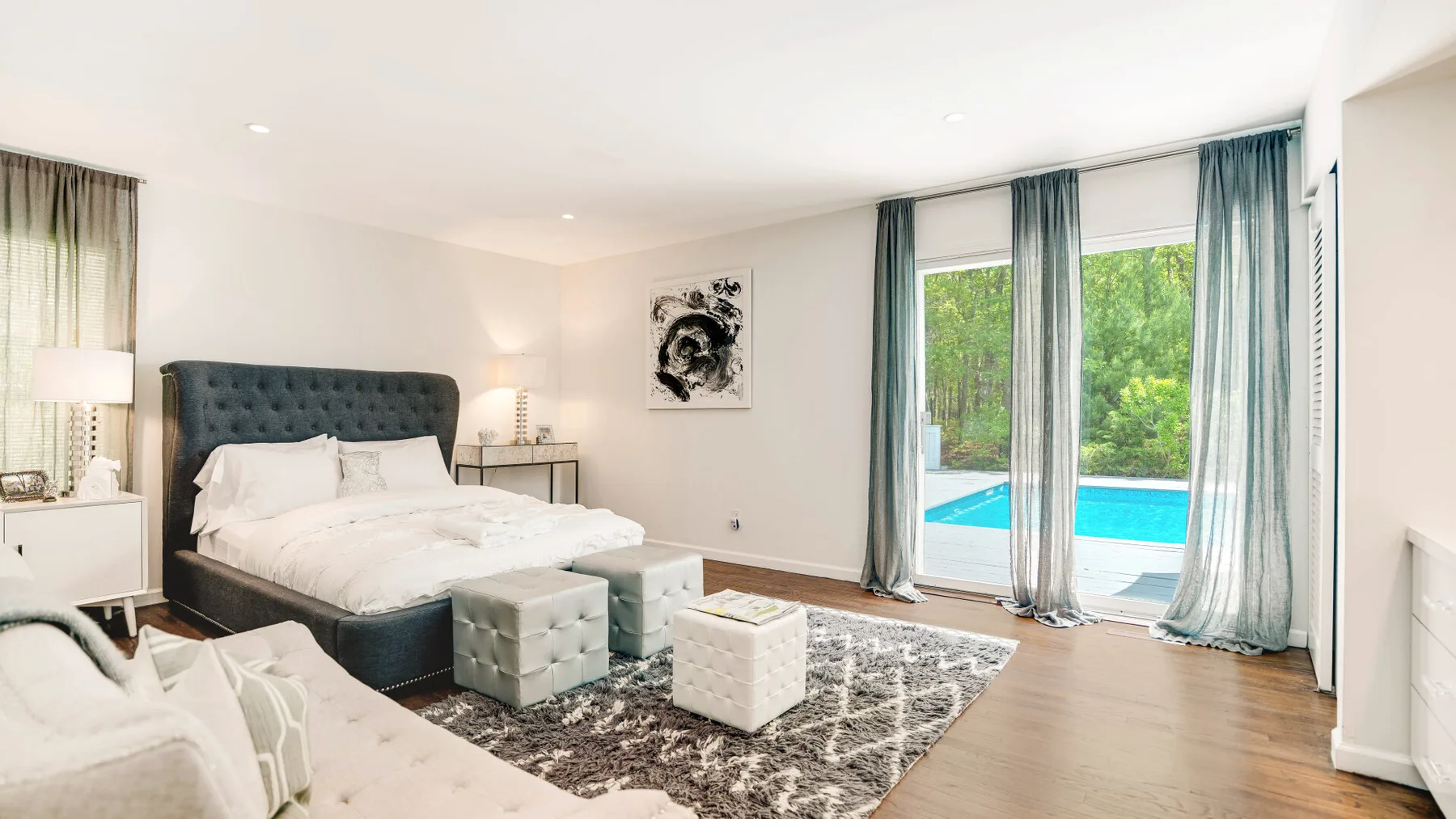 A bright bedroom with a bed, seating, and large windows overlooking a pool.