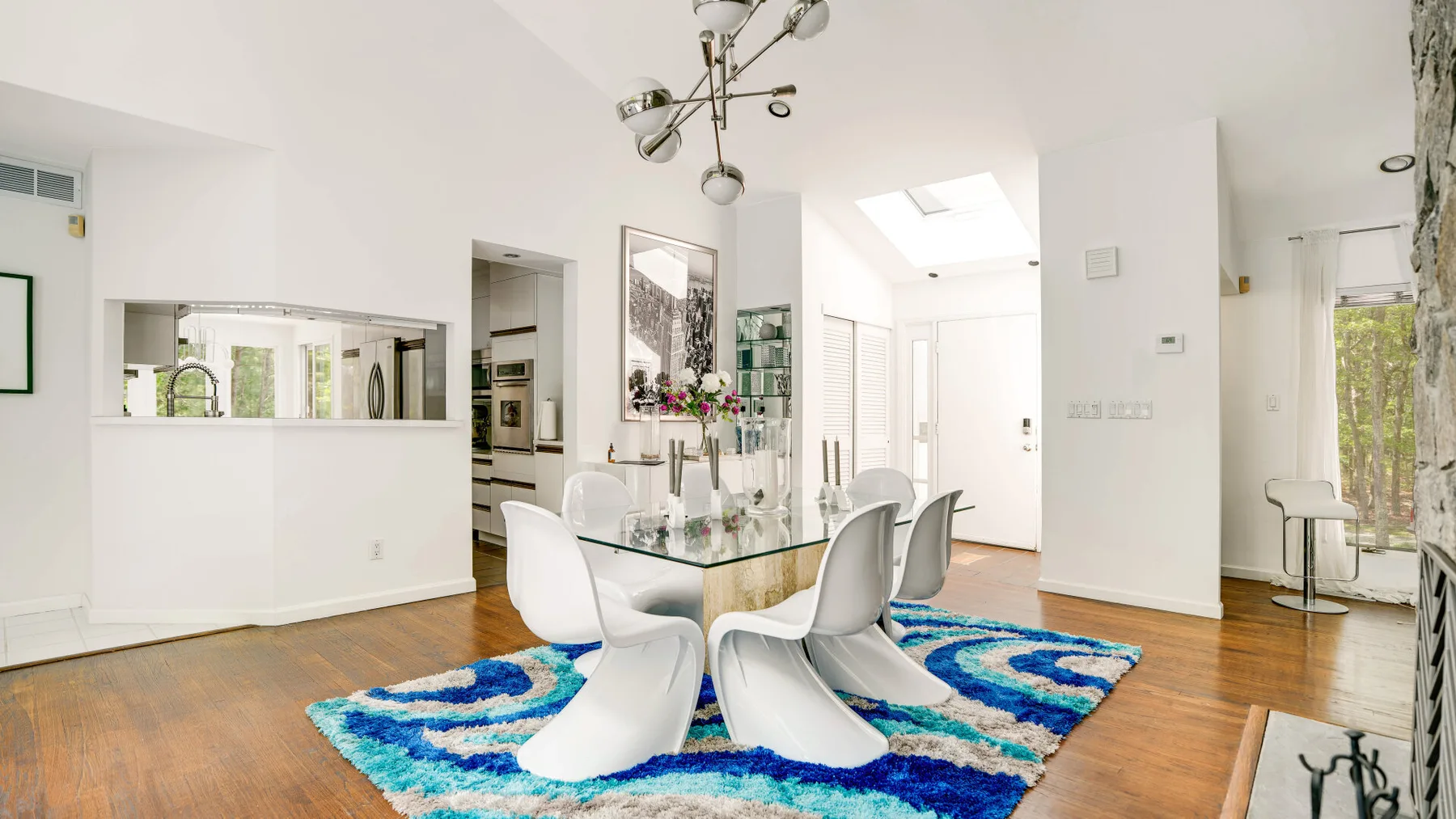 A bright dining area with a glass table and white seating above a colorful rug.