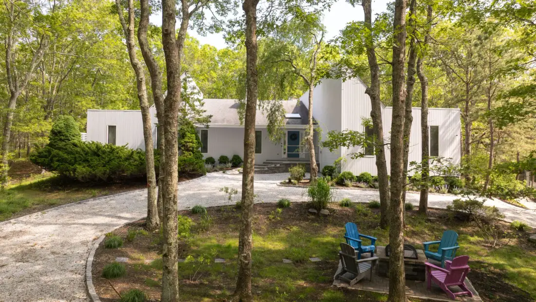 A modern house sits among trees, with outdoor seating beside the driveway.