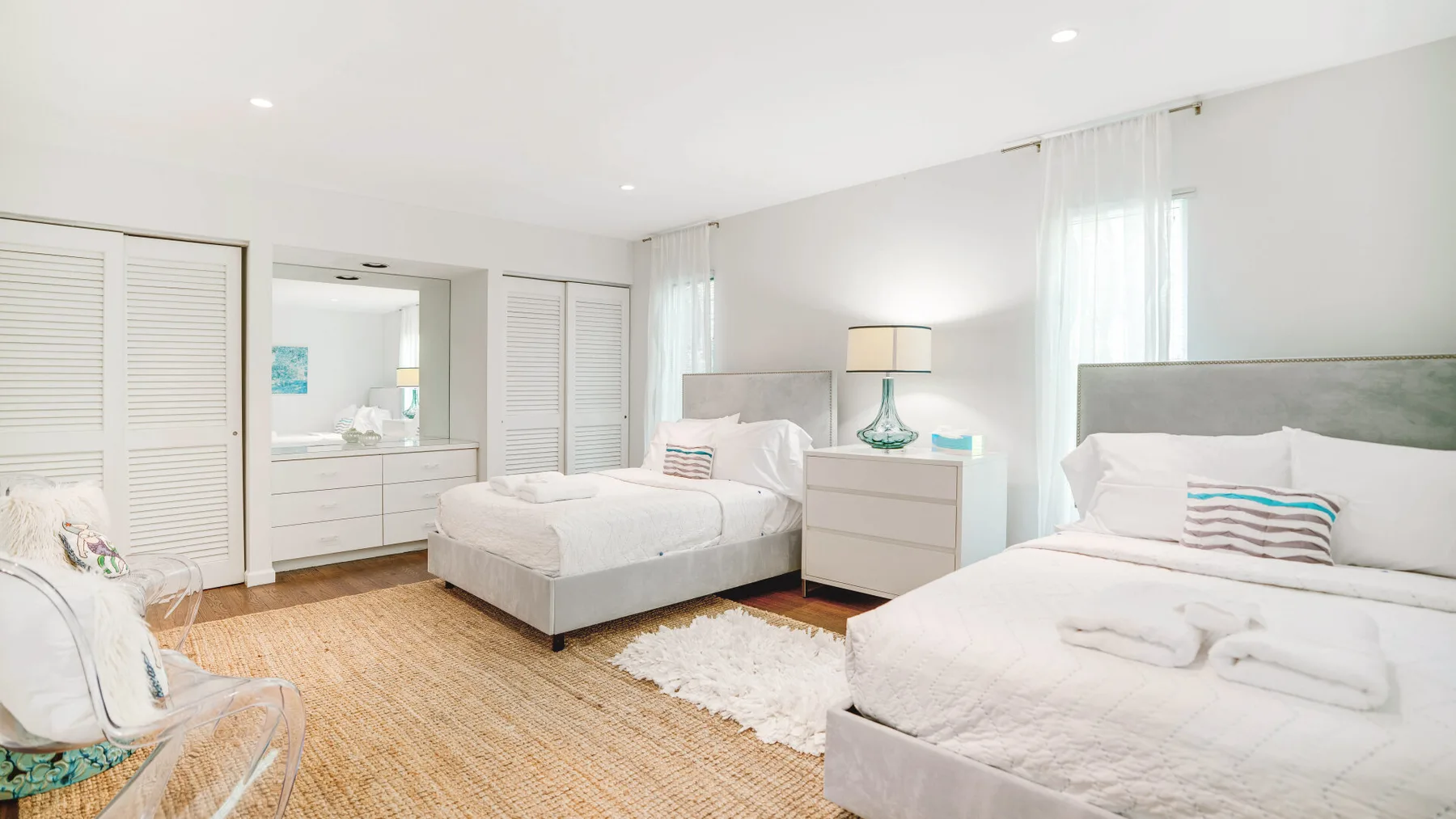 A bright bedroom with two beds, white linens, and a woven rug.