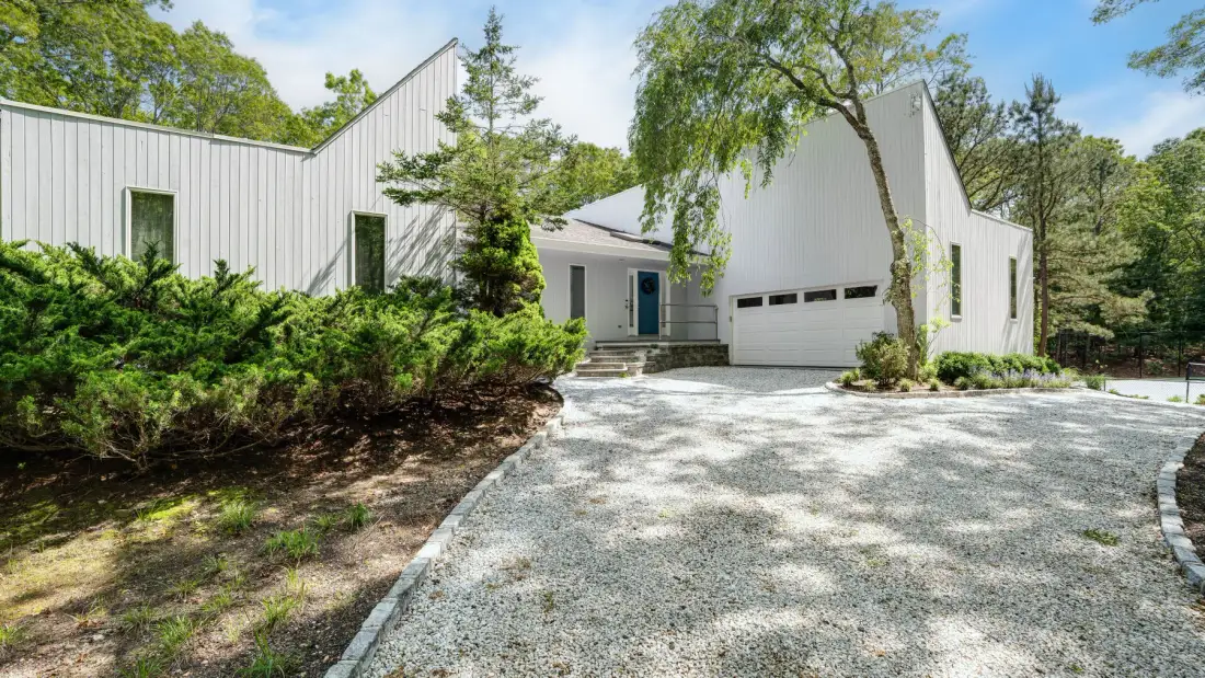Una casa blanca de estilo moderno se alza junto a una entrada de grava y vegetación.