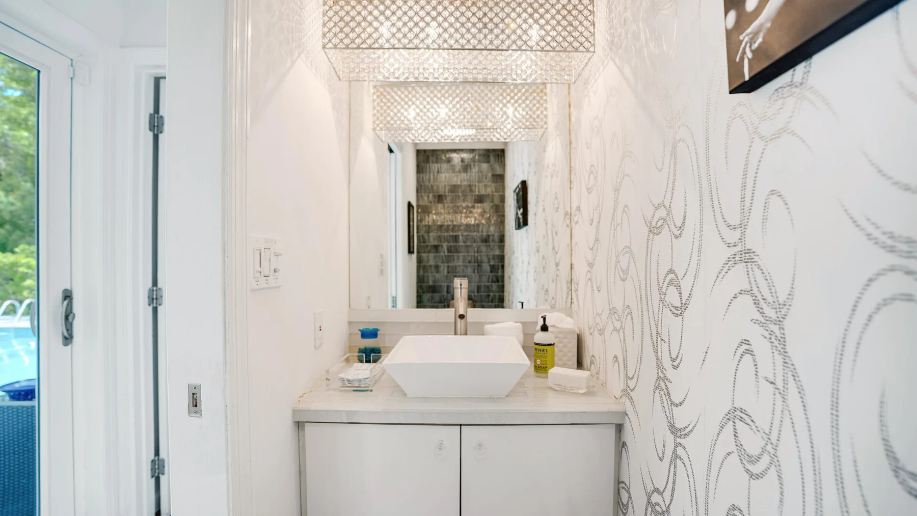 A bright bathroom vanity with a rectangular sink, mirror, and patterned wallpaper.