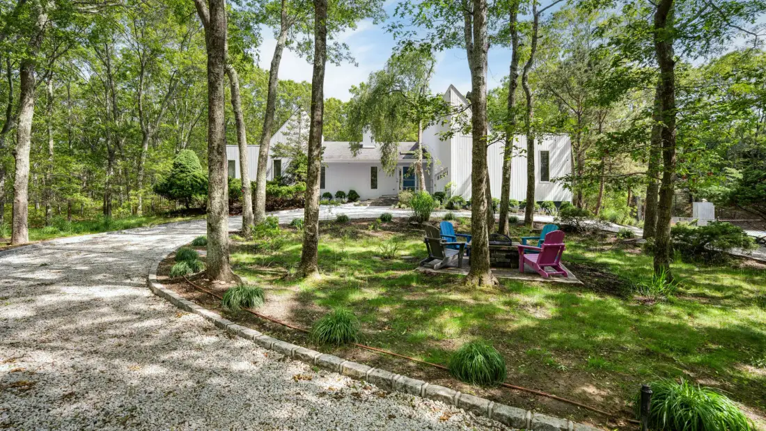 A white house sits among trees beside a curved gravel driveway and outdoor seating.
