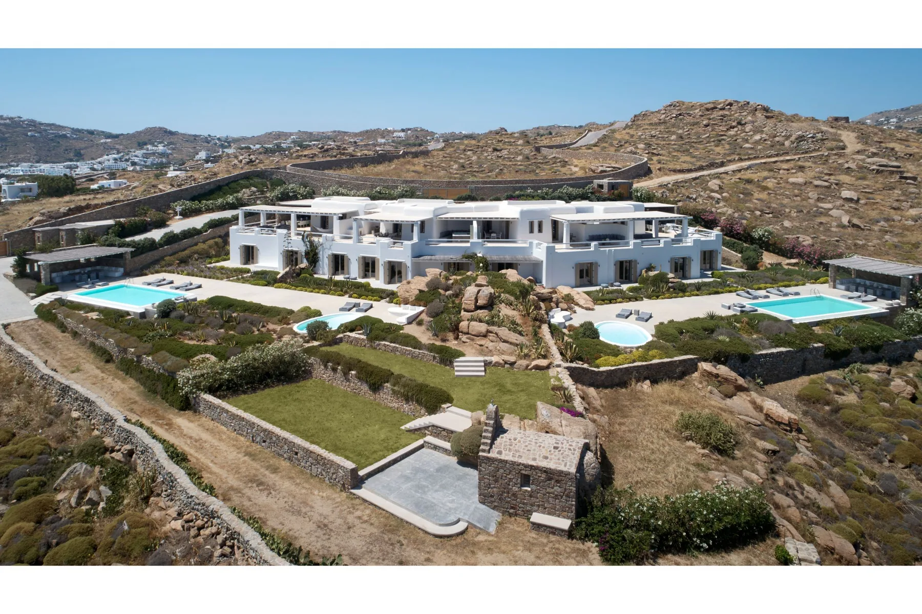 A large white villa with swimming pools sits among rocky hills.