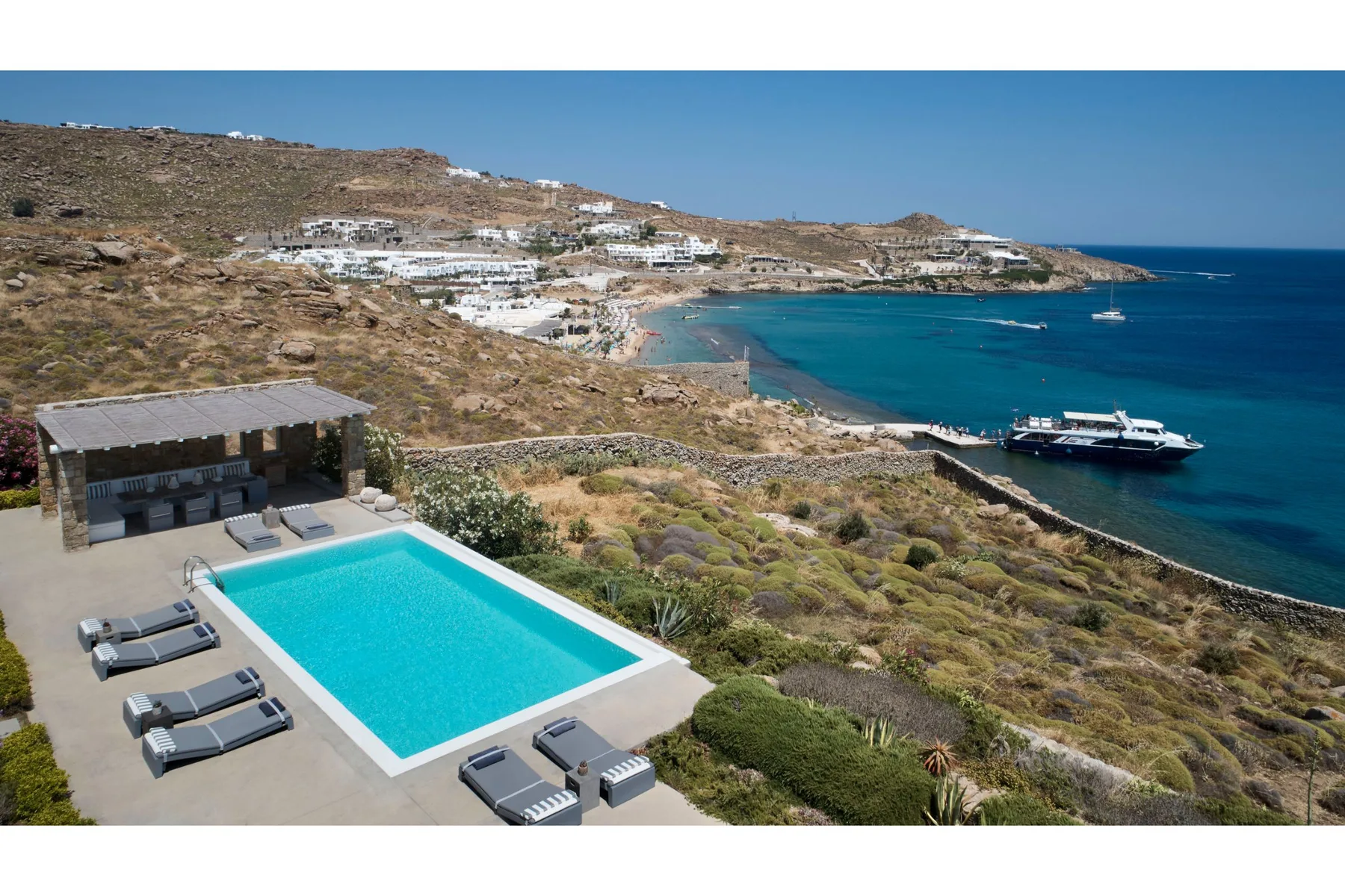 A seaside pool terrace overlooks a turquoise bay and coastal hillside.