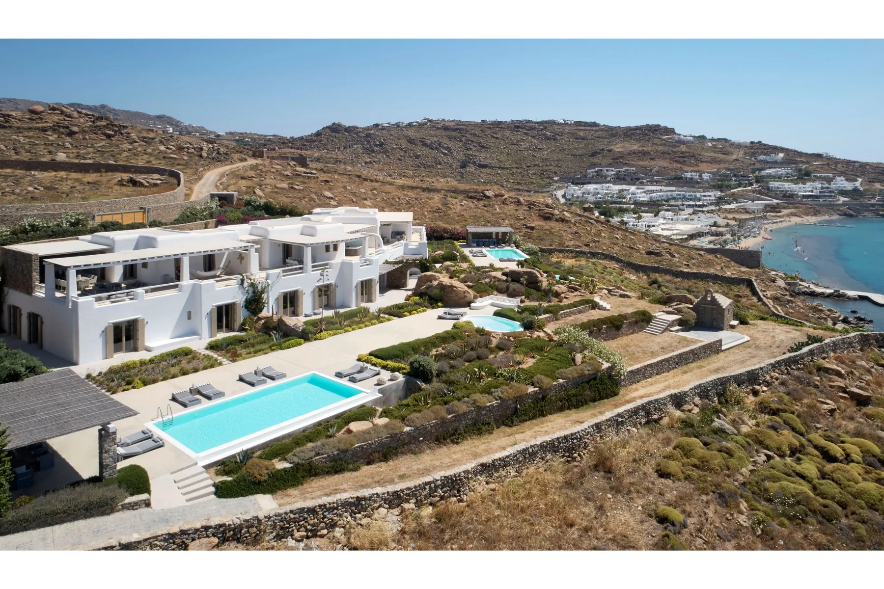 A white villa with swimming pools sits on a hillside above the coast.