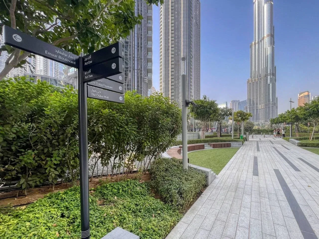 Una plaza peatonal ajardinada con caminos pavimentados, vegetación y edificios altos.