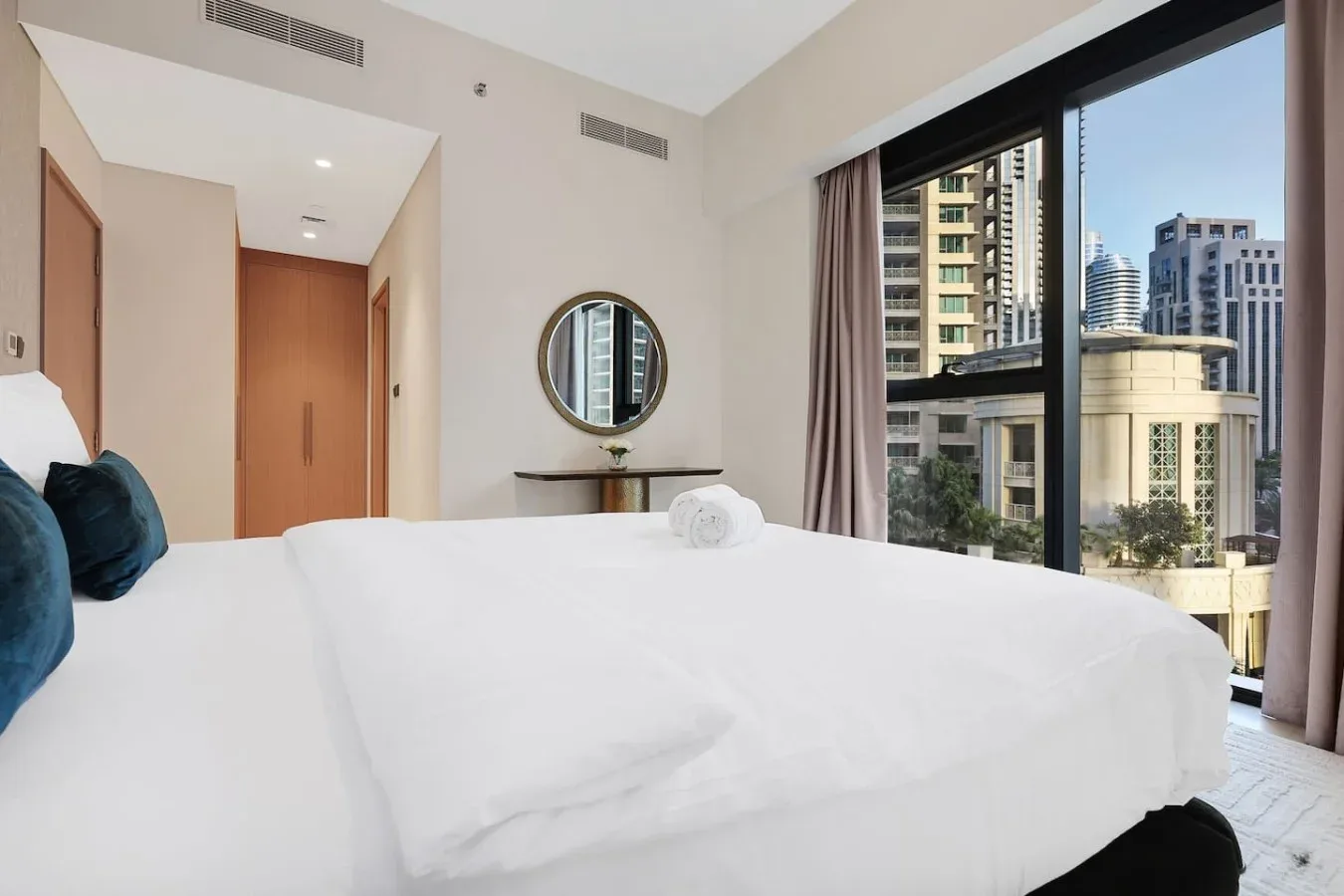 Un dormitorio luminoso con una cama bien tendida y grandes ventanas con vistas a edificios cercanos.
