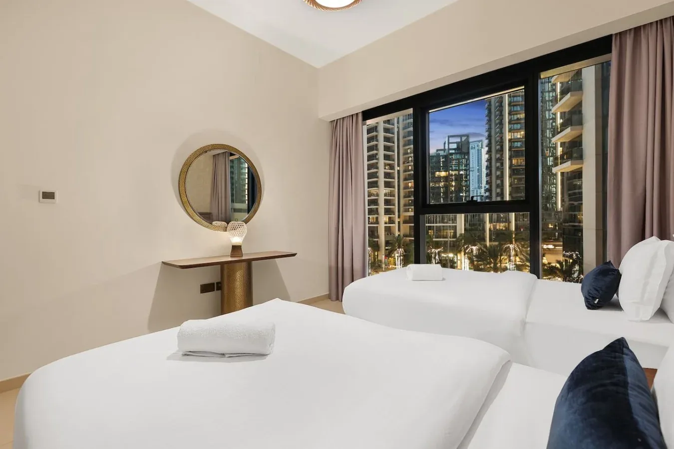 Un dormitorio luminoso con ropa de cama blanca y grandes ventanas con vistas a edificios cercanos.