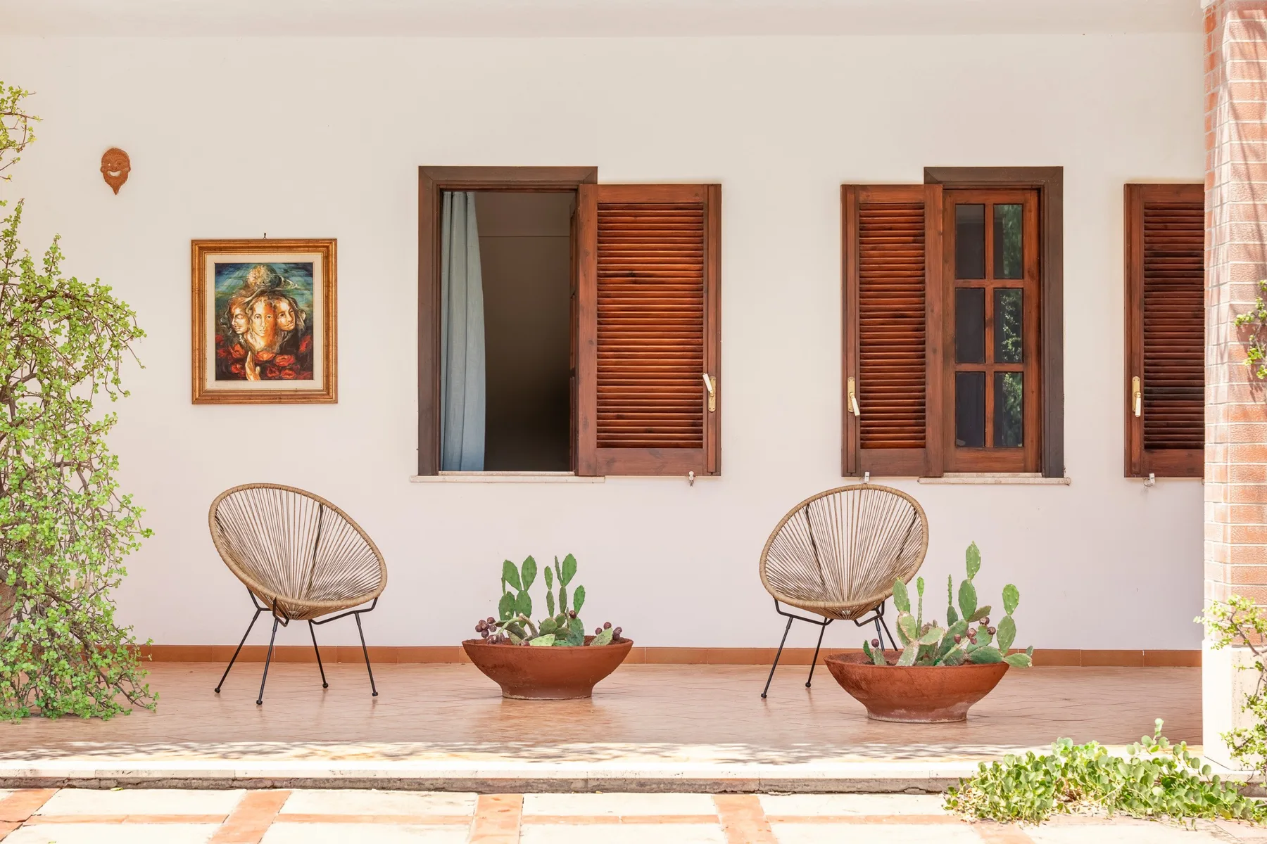 Un patio de paredes blancas con contraventanas de madera, asientos tejidos y plantas en macetas.