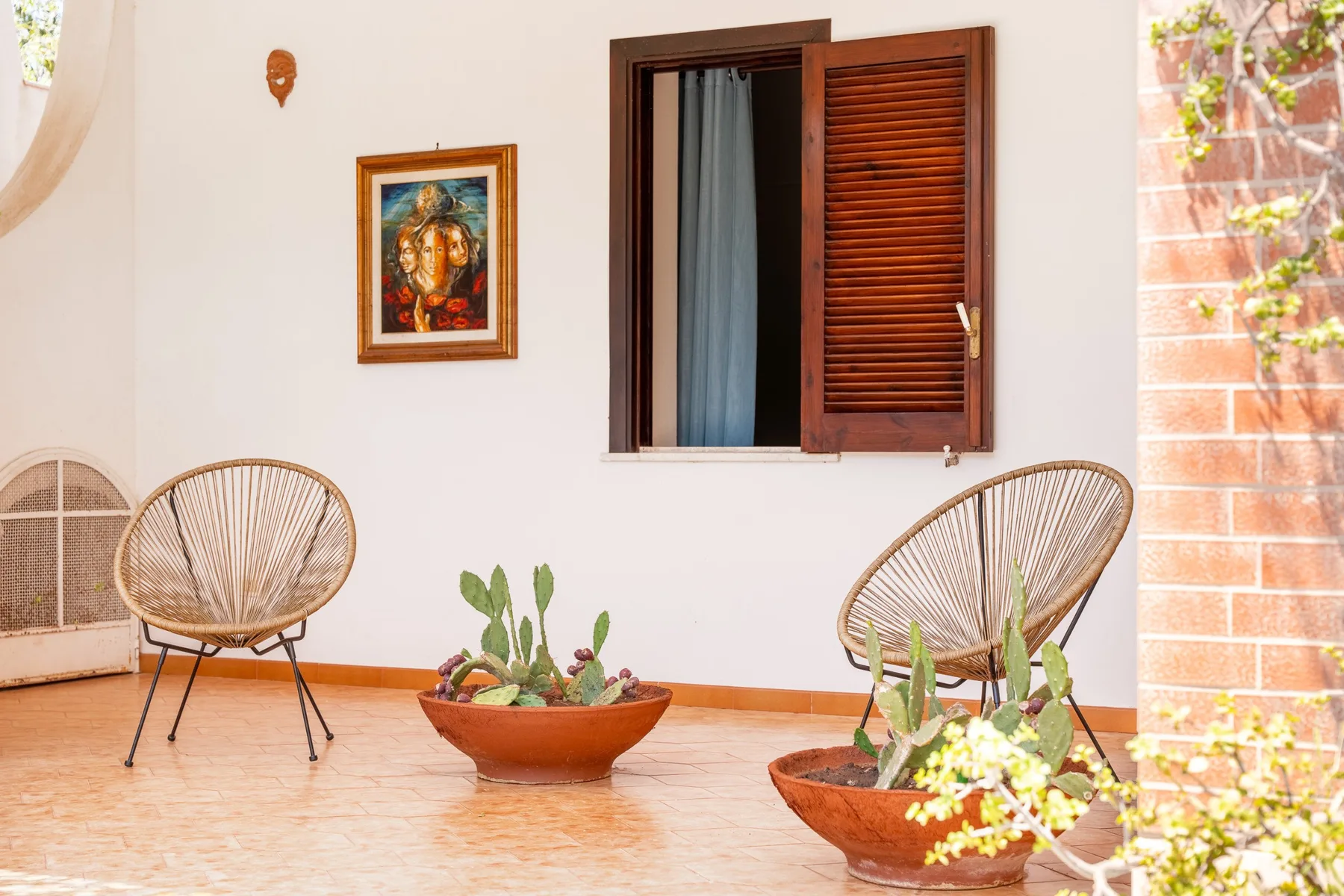 Un patio luminoso con sillas tejidas y plantas en macetas junto a una ventana abierta.