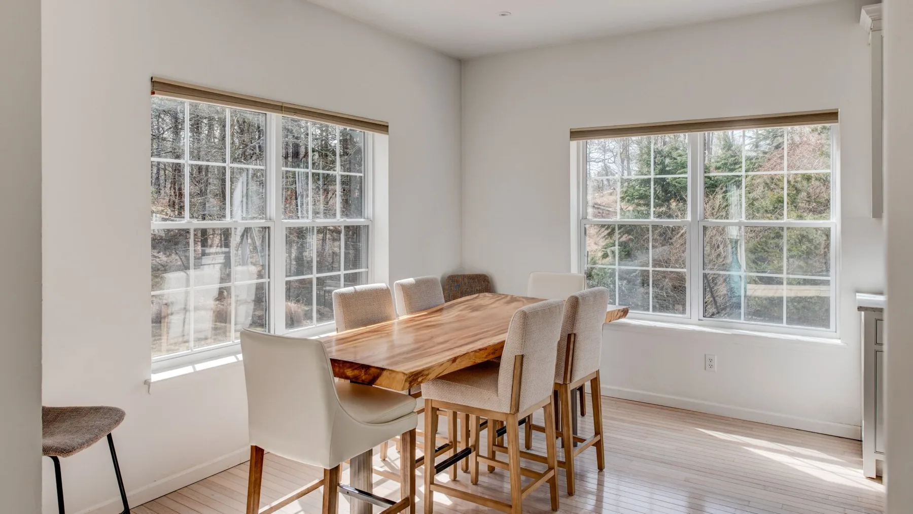 A dining area with a wooden table and upholstered seating beside two large windows.