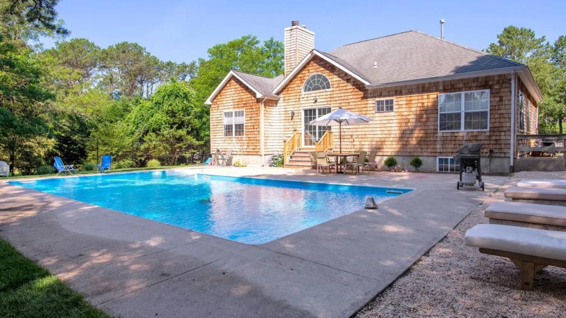 A backyard pool beside a house, with patio seating and trees.
