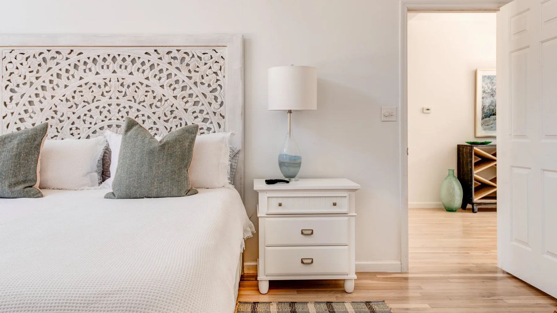 A bedroom with a white bed, decorative headboard, and bedside table beside an open doorway.