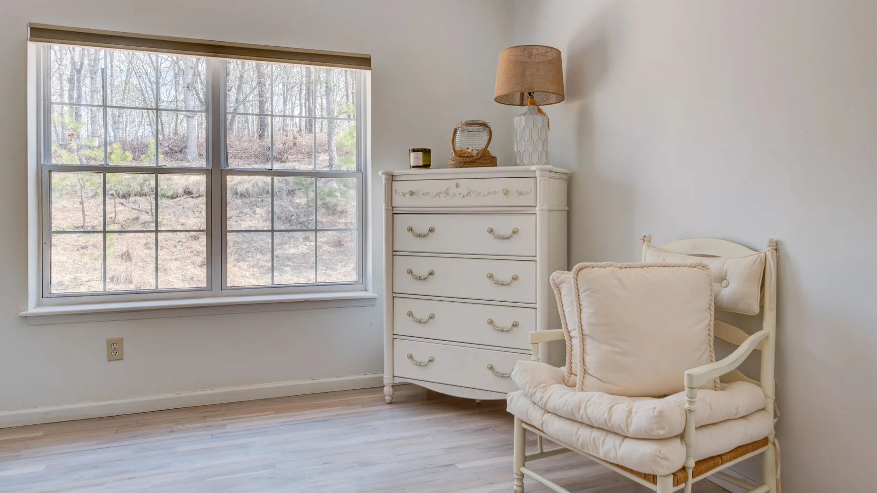 A bright bedroom sitting area with a cushioned chair beside a dresser and large window.