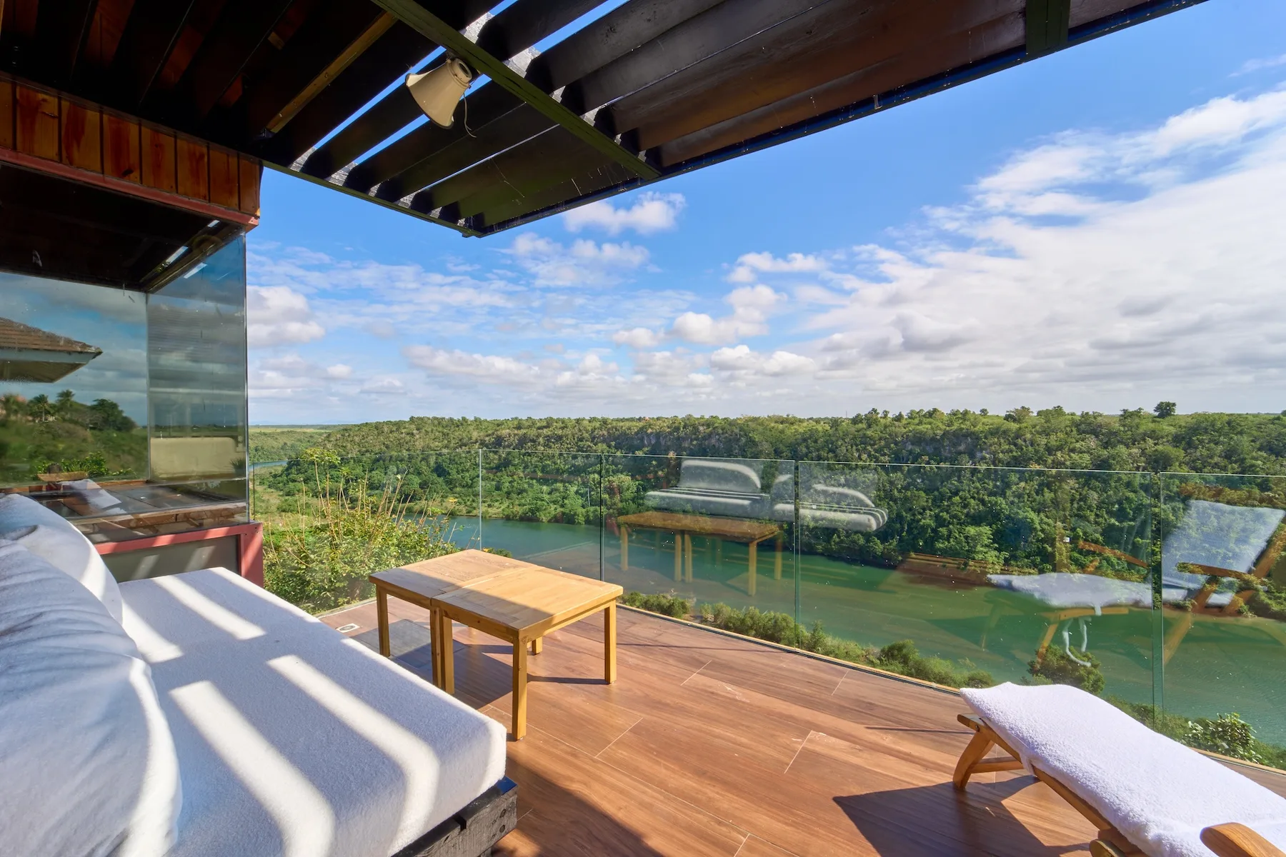 Una terraza abierta de madera con asientos de descanso mira hacia un paisaje verde bajo un cielo parcialmente nublado.