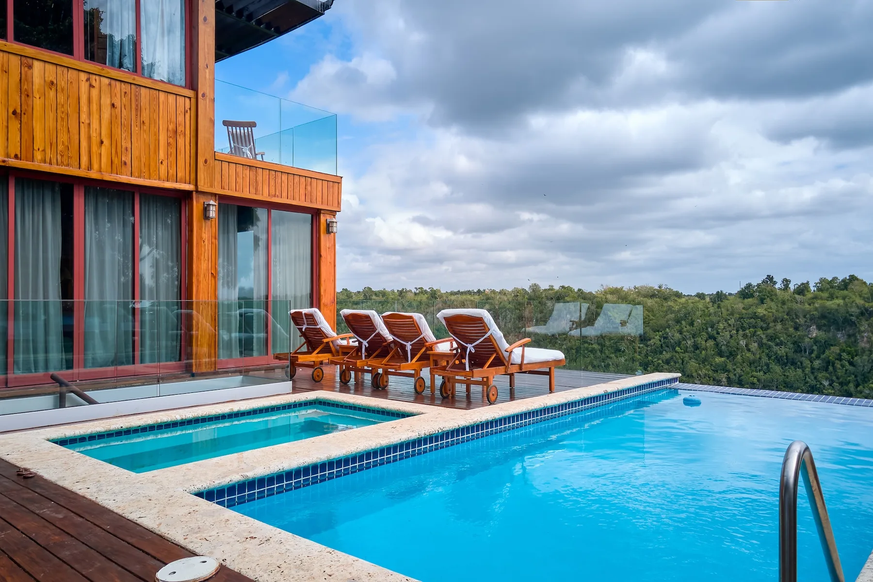 Outdoor pool beside a modern building, with lounge seating overlooking a wooded landscape.