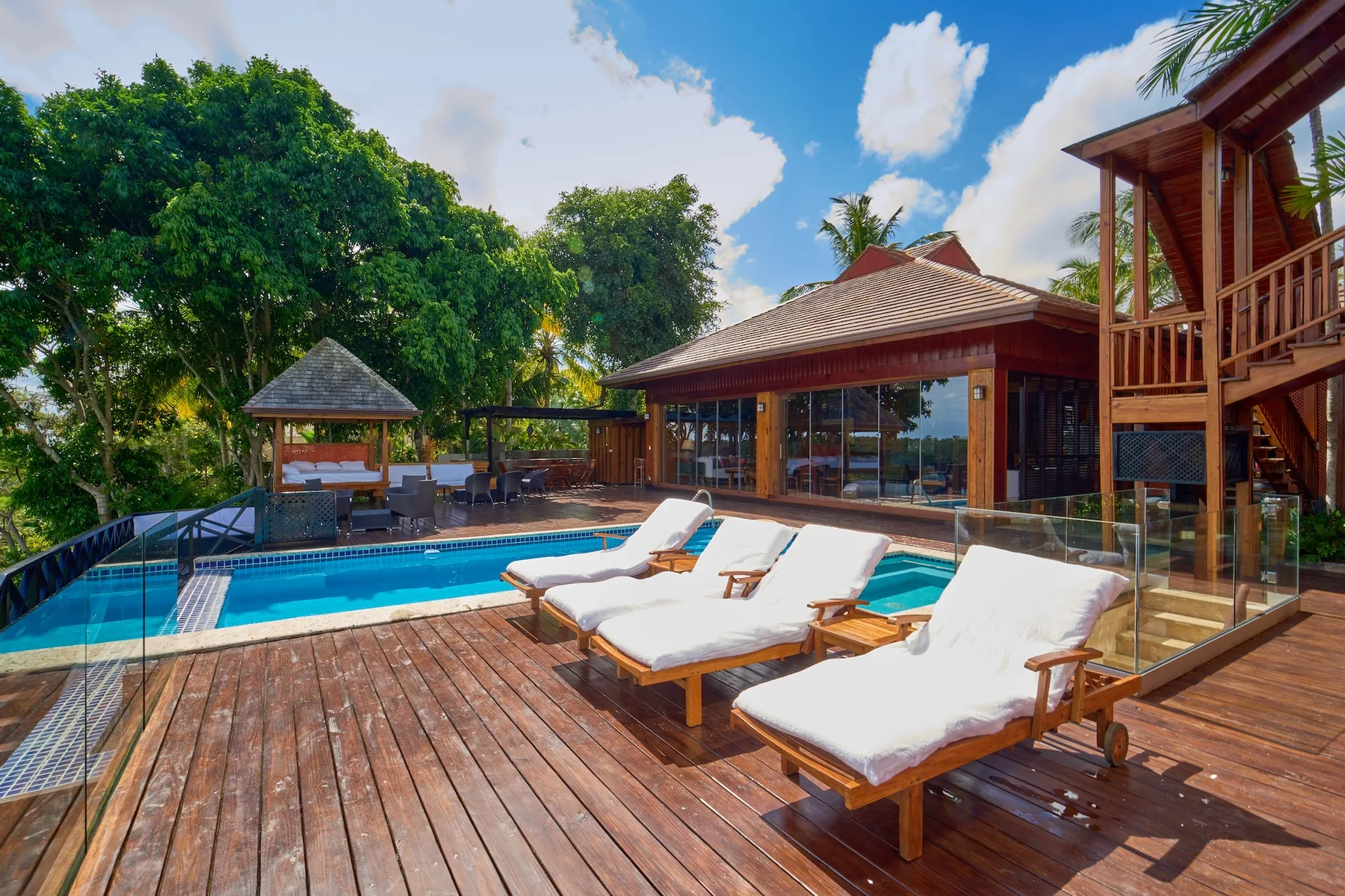 A poolside patio with lounge seating beside a wooden building and leafy trees.