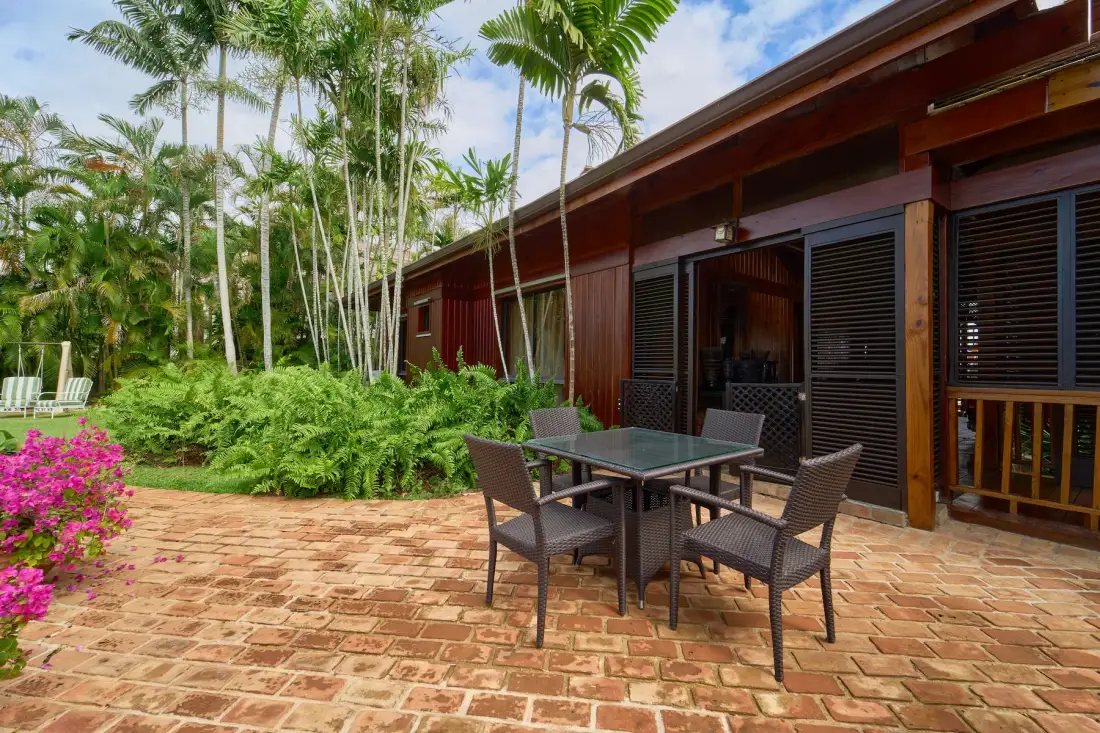 Un comedor en un patio junto a un edificio de madera oscura, rodeado de plantas tropicales.