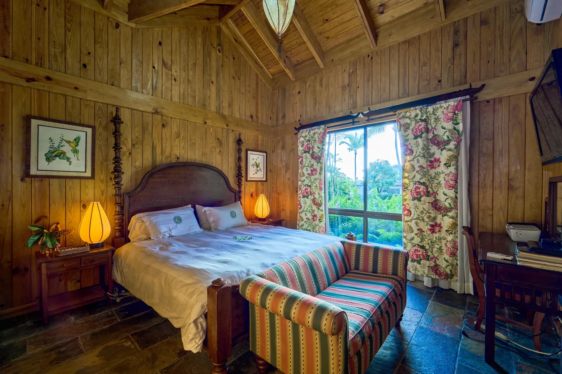 Un dormitorio revestido de madera tiene una cama frente a una ventana grande con cortinas florales.