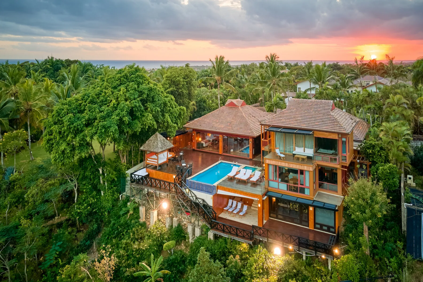 Una casa tropical con piscina está rodeada de vegetación al atardecer.