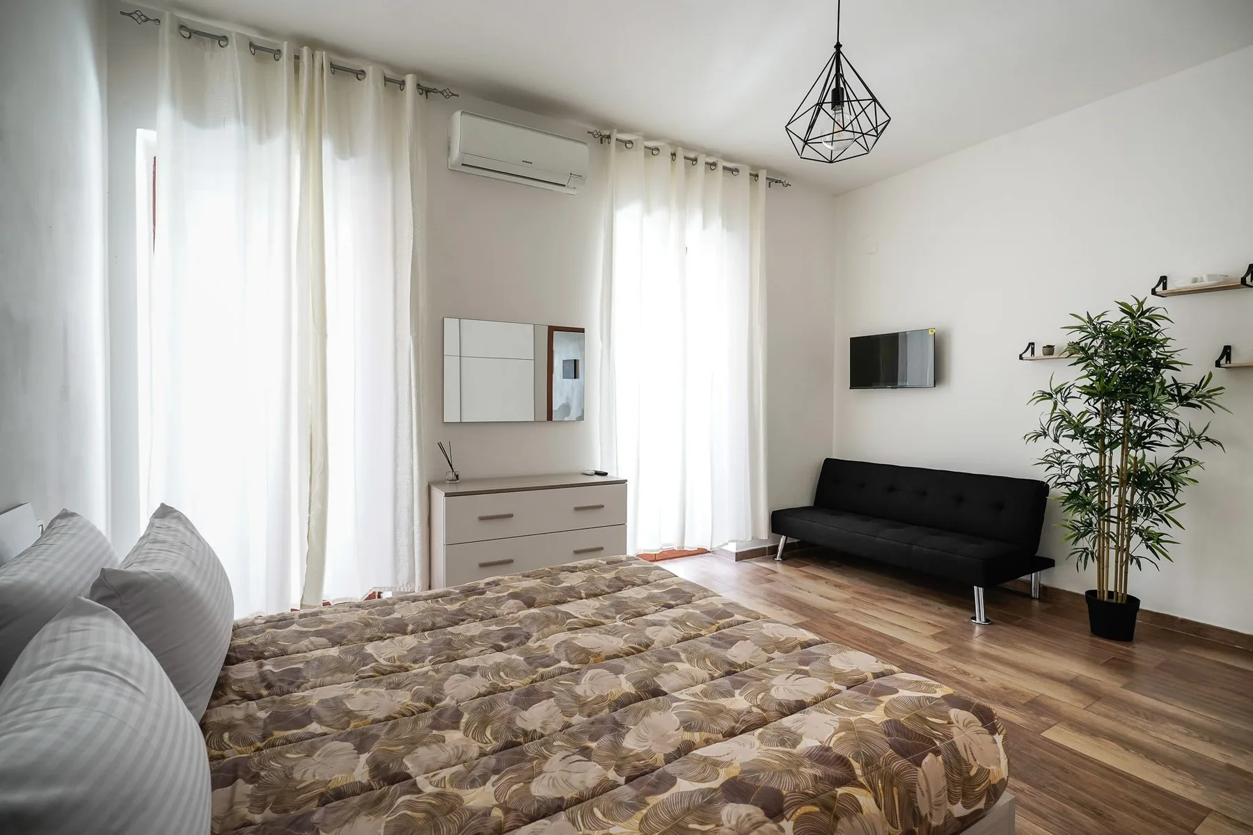 Un dormitorio luminoso con una colcha estampada, asientos negros y ventanas altas con cortinas.