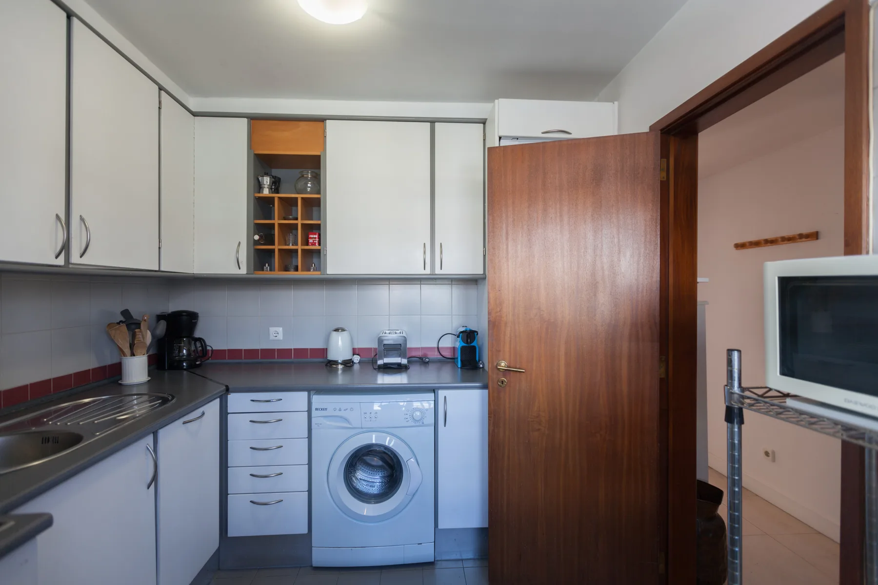 Una cocina compacta con armarios blancos, una lavadora y una puerta de madera abierta.