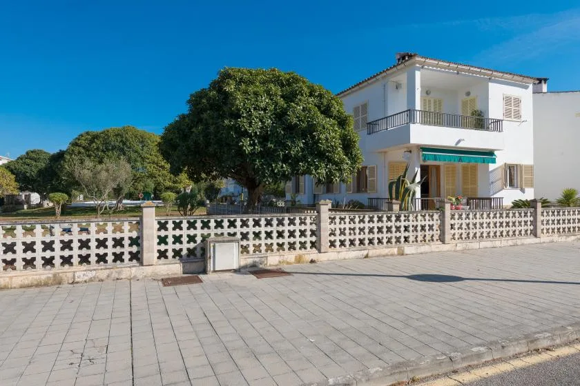 A white two-story house stands behind a patterned fence, with trees in the yard.