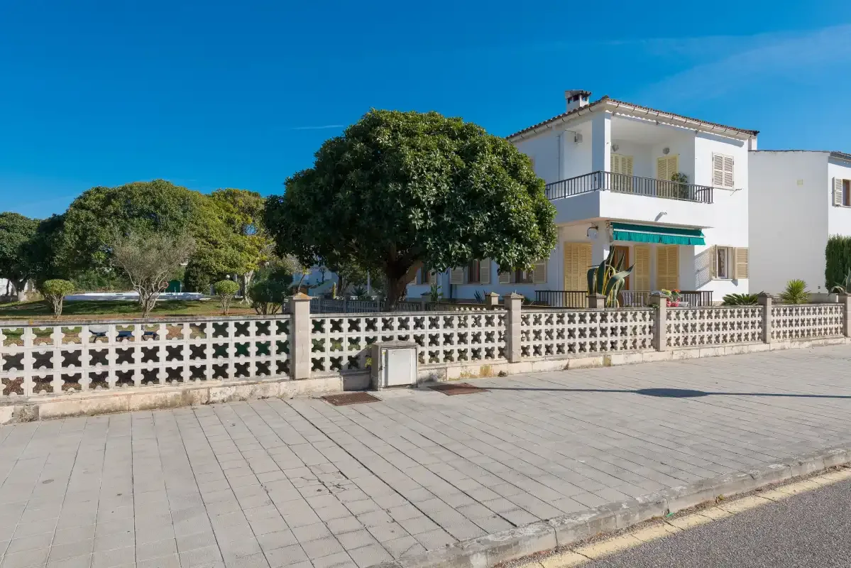 A white two-story house stands behind a patterned low wall beside a paved street.