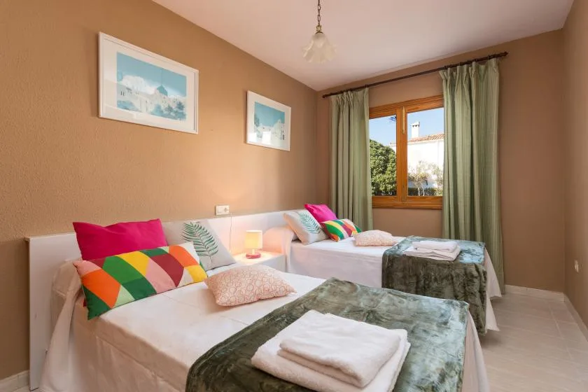 A bedroom with two beds, colorful pillows, and a window with green curtains.