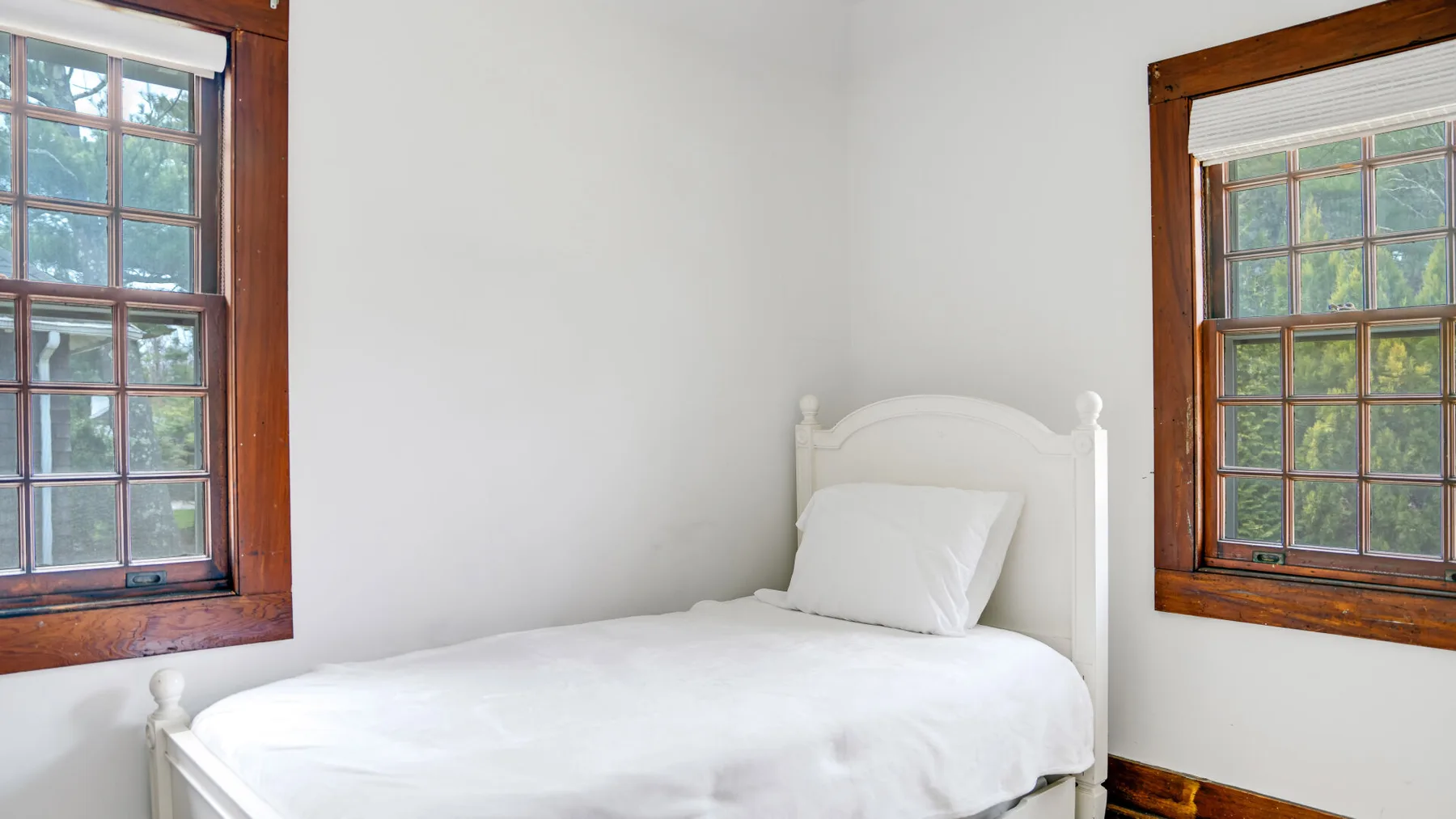 Un dormitorio con una cama blanca entre dos ventanas con marcos de madera.