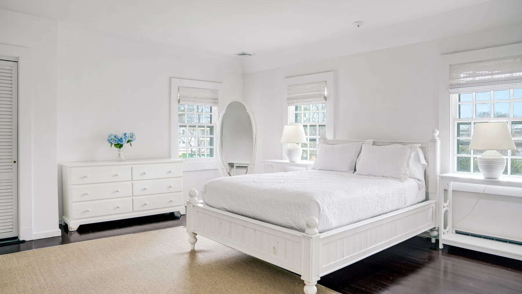 Un dormitorio luminoso con una cama blanca, una cómoda y varias ventanas.