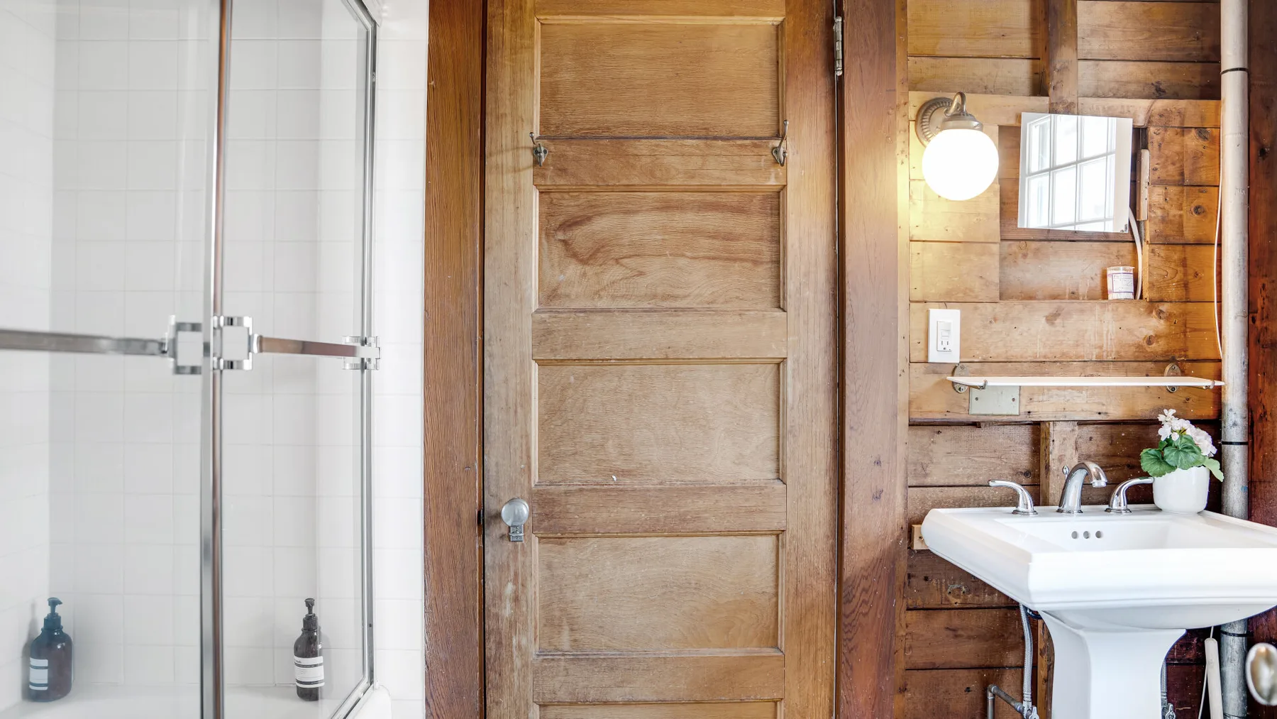 Un baño con ducha de vidrio, puerta de madera y lavabo con pedestal.