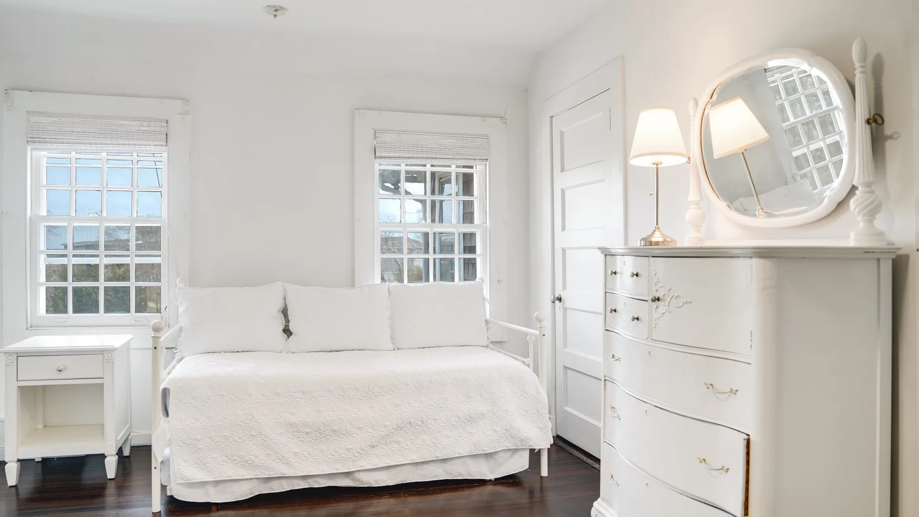 Un dormitorio luminoso con una cama blanca, una mesita de noche y una cómoda.
