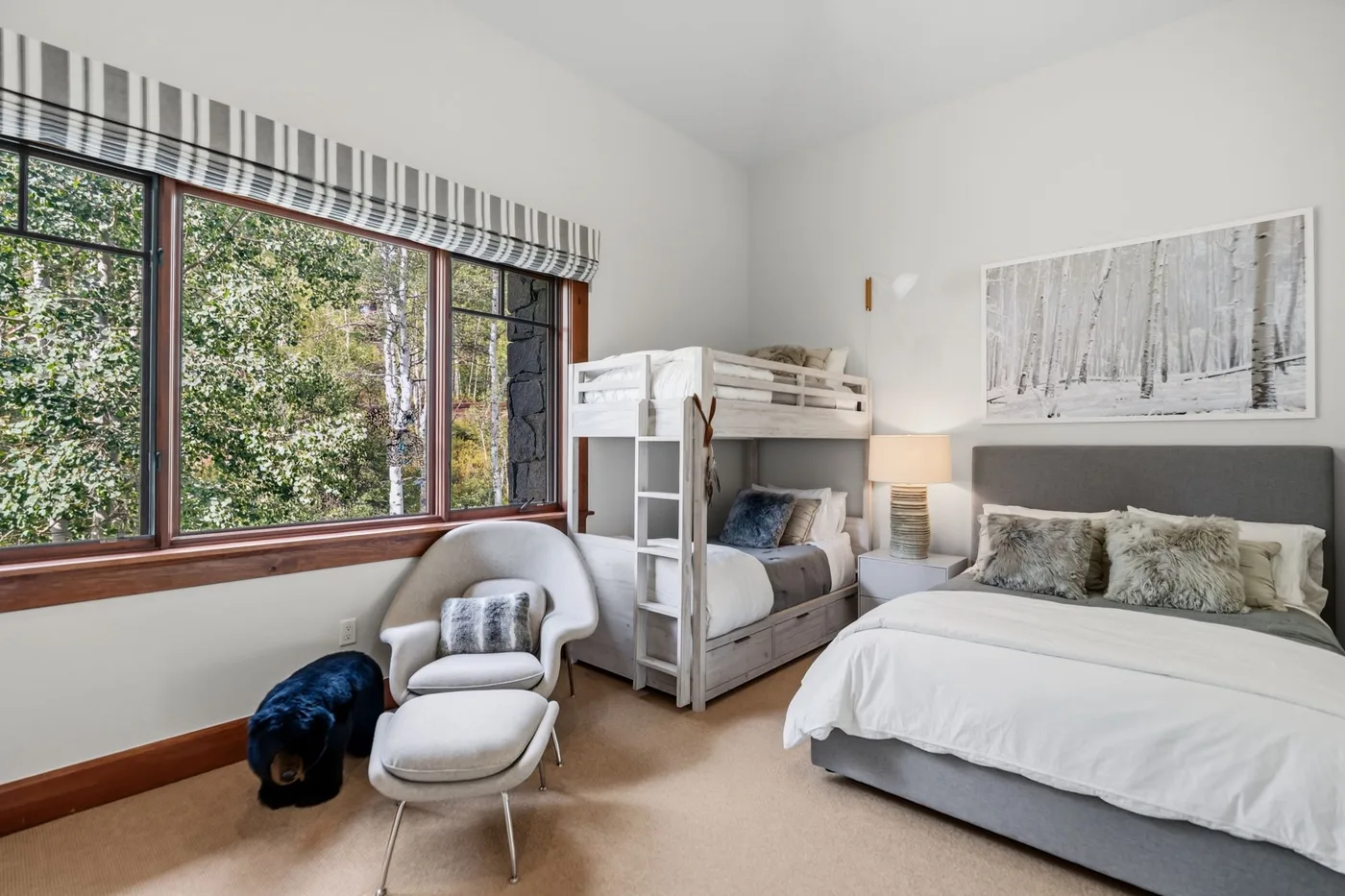 A bedroom with a bunk bed, a larger bed, and windows overlooking trees.