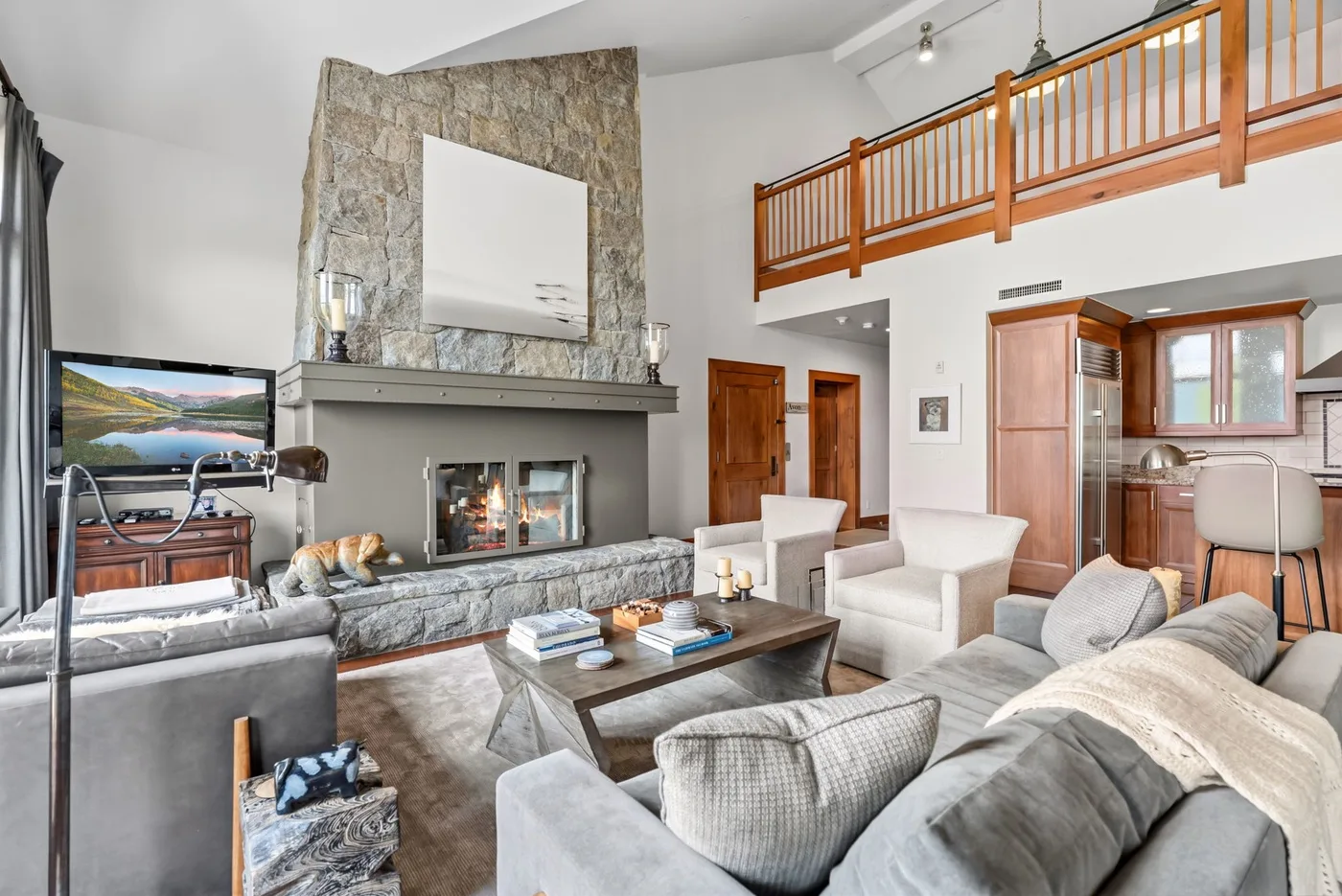 A spacious living room with a stone fireplace, seating, and a wooden balcony railing.