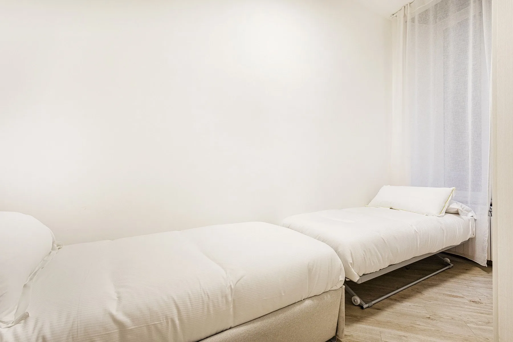 Un dormitorio luminoso con dos camas y cortinas translúcidas junto a la ventana.