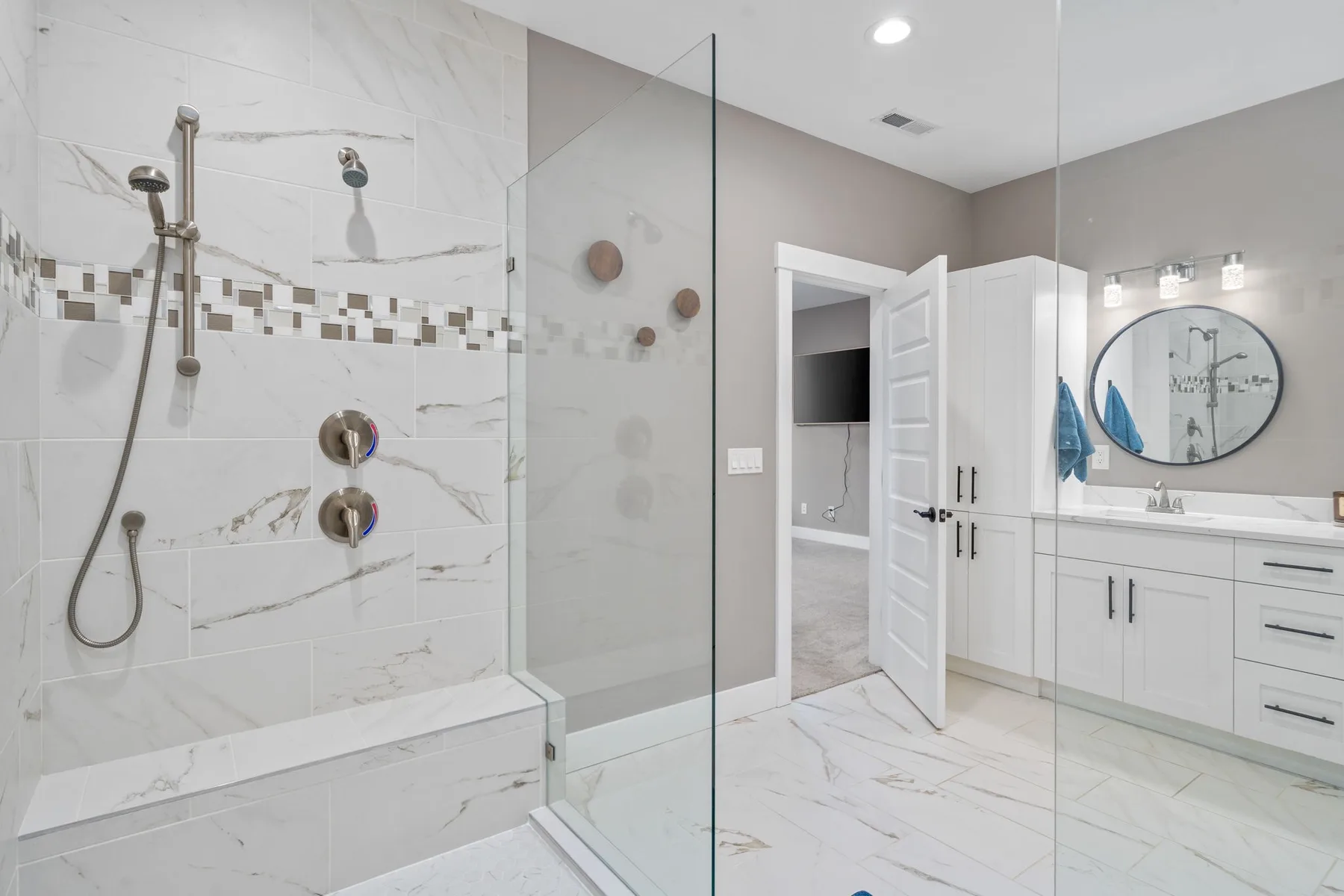 Un baño luminoso con ducha de vidrio, tocador blanco y espejo redondo.