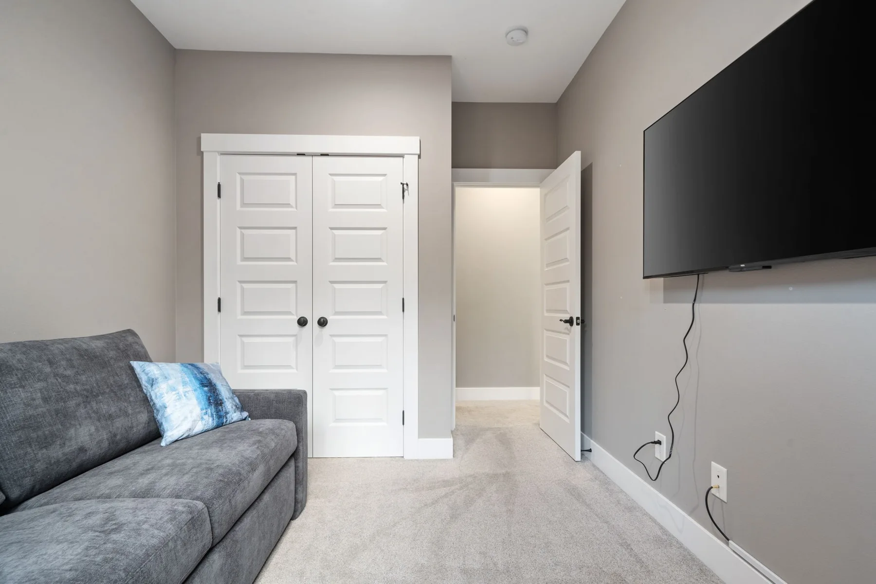 Sala alfombrada con un sofá gris, un televisor montado en la pared y una puerta abierta.