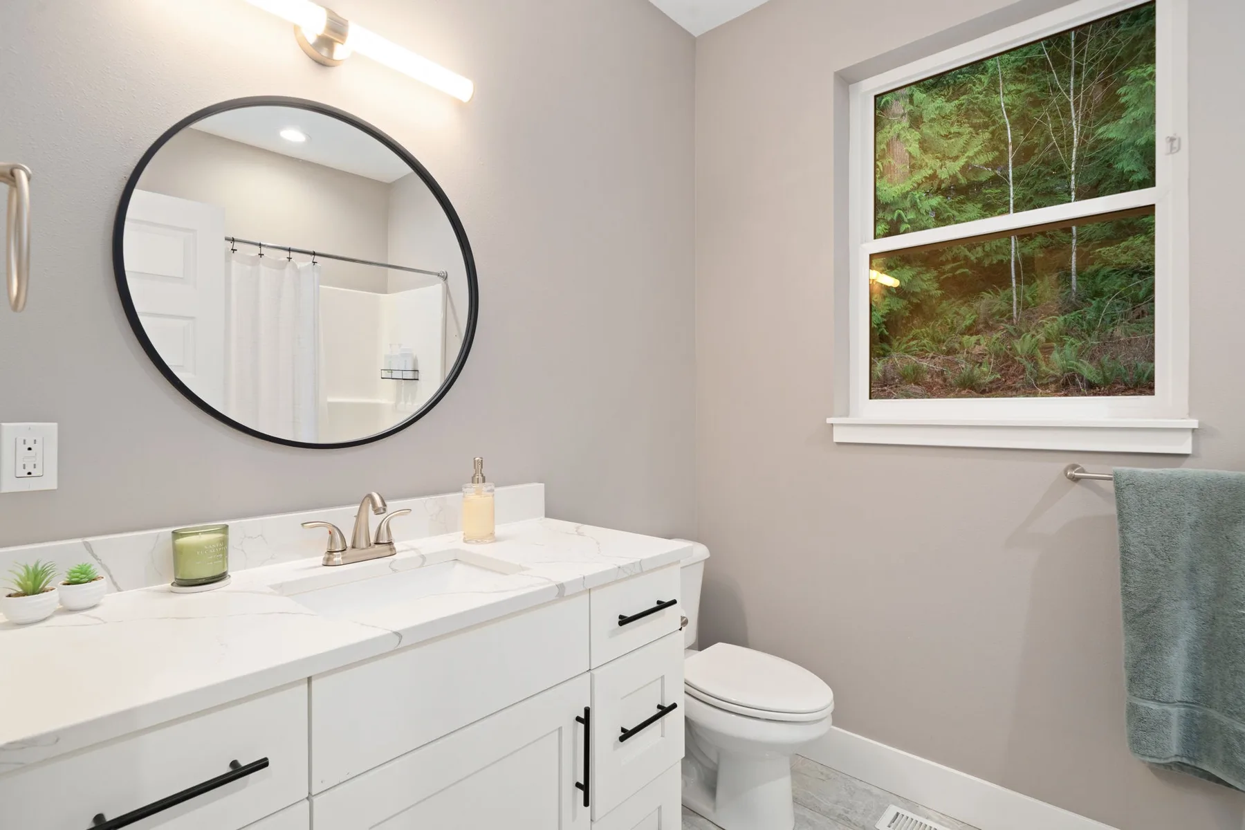 Un baño luminoso con un tocador blanco, un espejo redondo y una ventana.