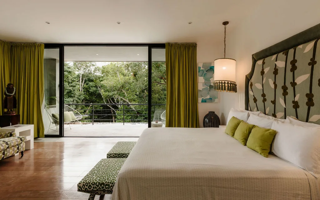 A bedroom with a large bed facing glass doors that open onto a leafy balcony.
