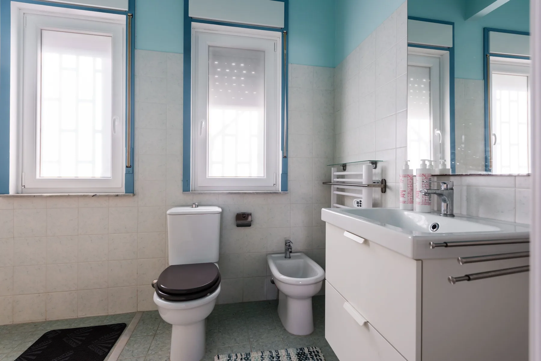 Un baño luminoso con inodoro, bidé y lavabo bajo ventanas altas.